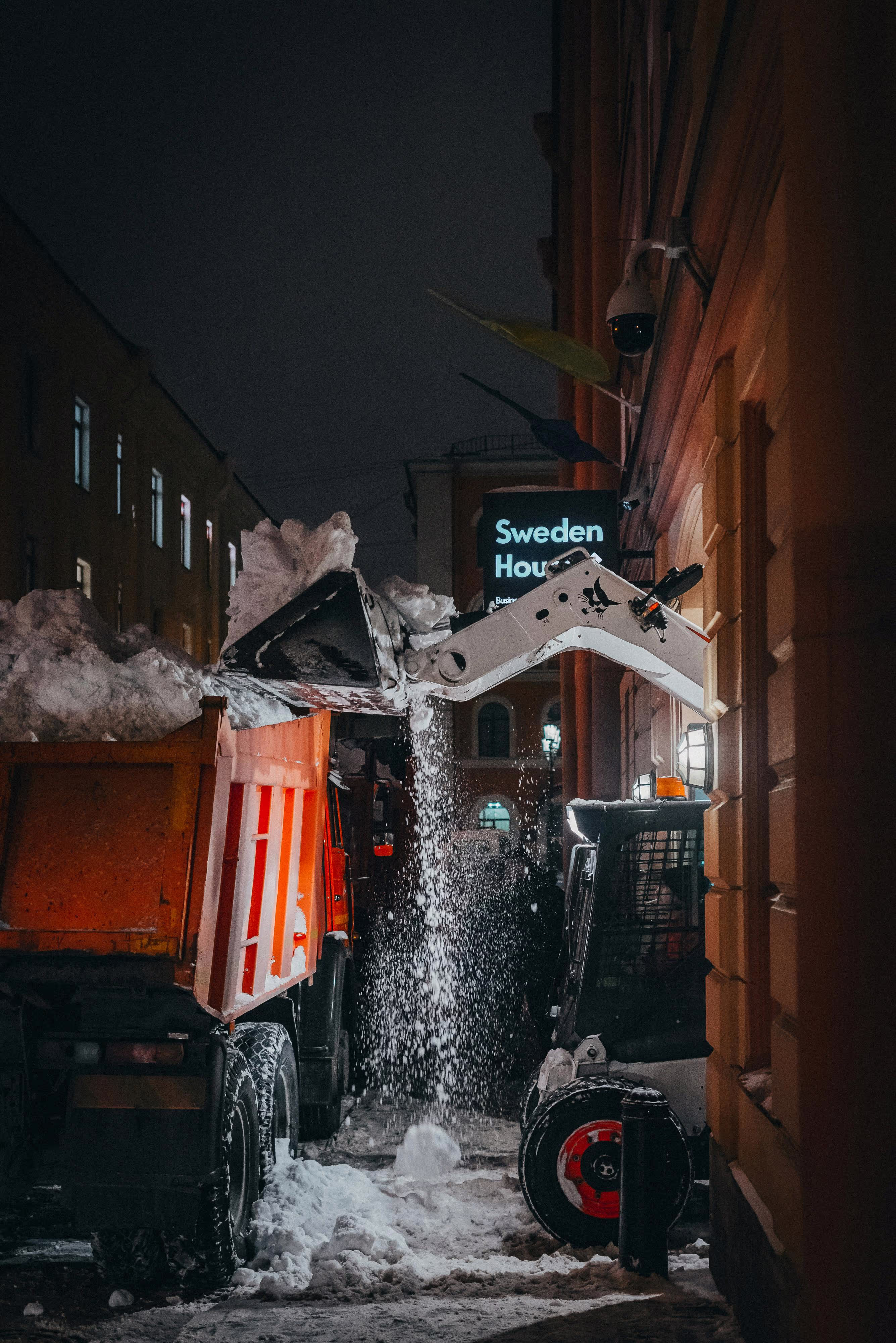 Ein Bagger kippt Schnee in einen vor ihm stehenden Kipp-Lkw.